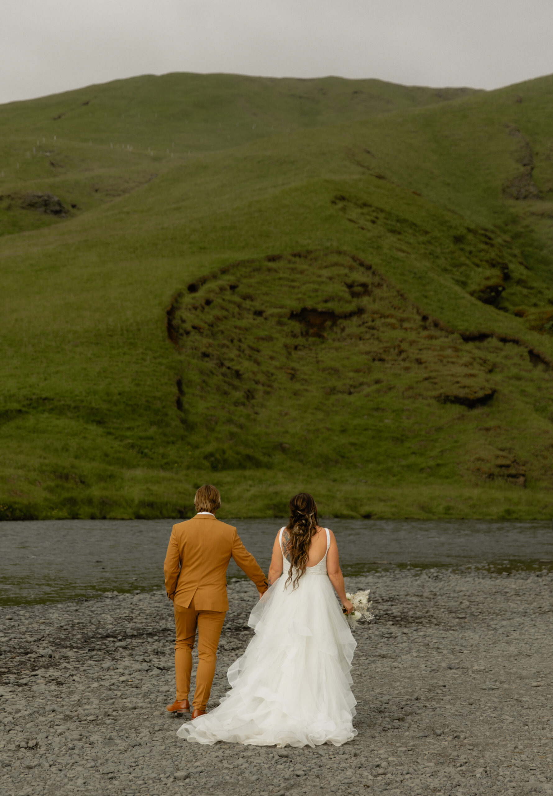 Bride and groom walking together beneath green cliffs near Skógafoss waterfall in Iceland