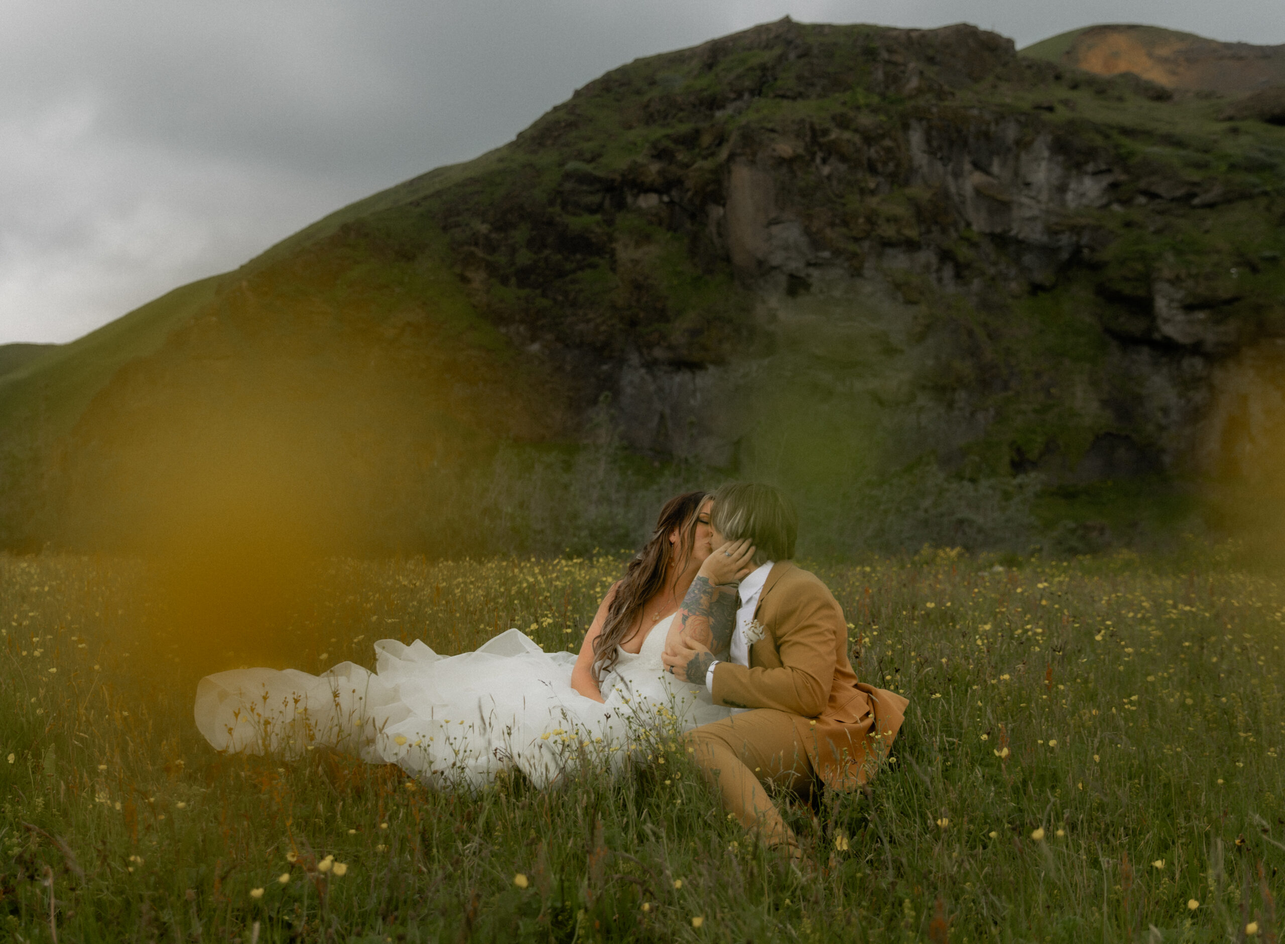 Bride and groom kissing in wildflower field beneath Icelandic cliffs