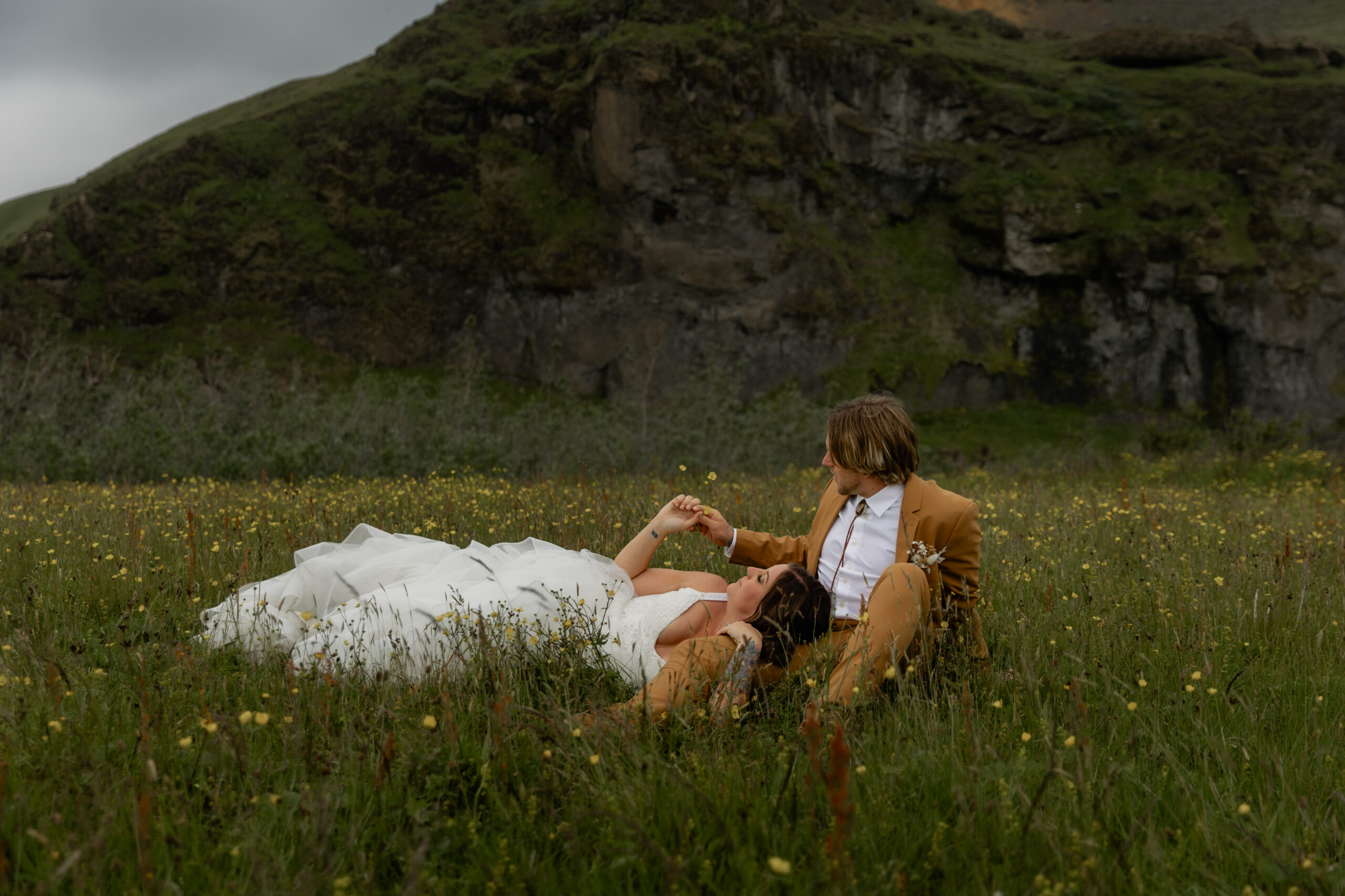 Bride and groom relaxing together in Iceland wildflower field beneath moss-covered cliffs
